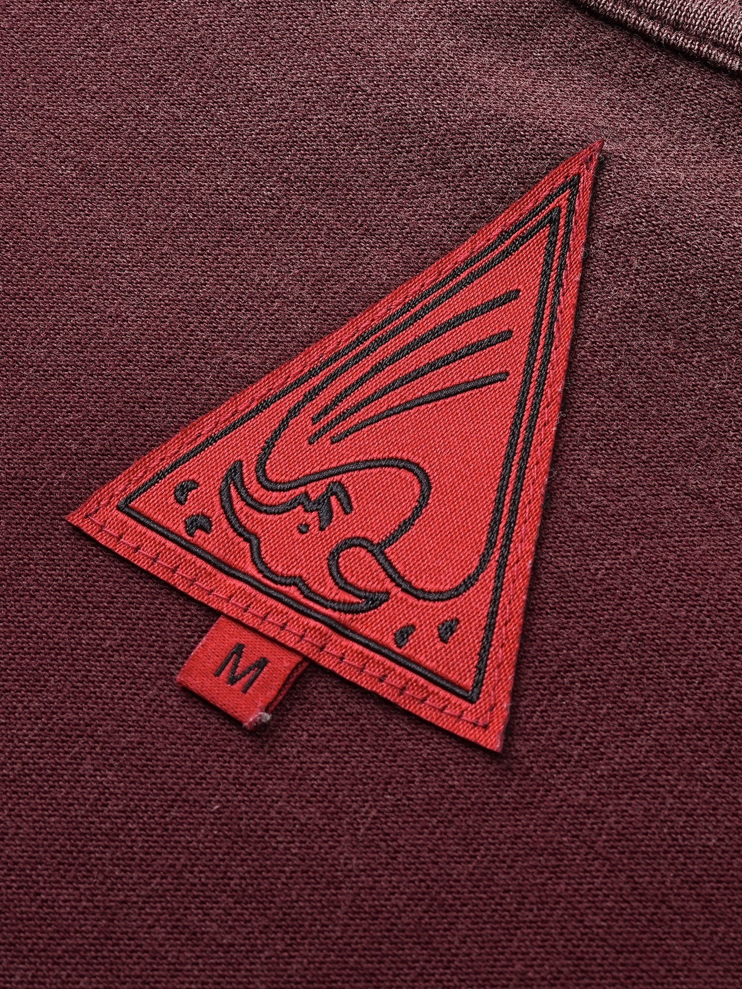 Close-up of the red triangle logo patch on TBHNP burgundy Wraith Sorcerer tee — men's streetwear detail