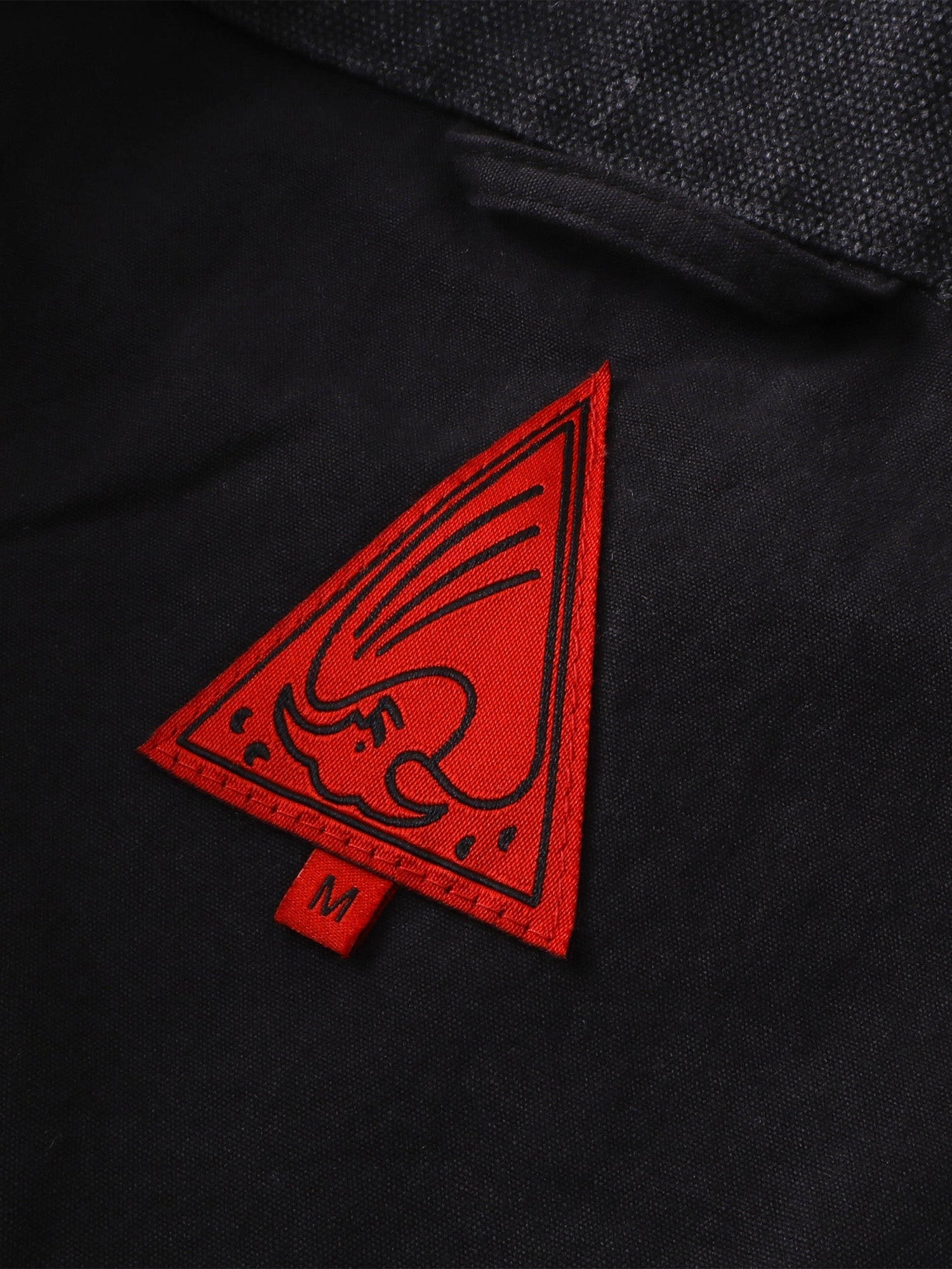 Close-up of the red triangle logo patch on TBHNP canvas moto jacket — men's streetwear detail